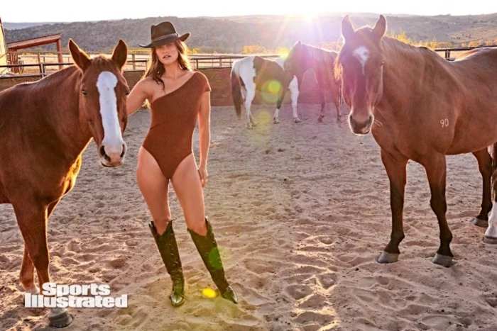 Myla Dalbesio poses next to two brown horses in a brown one-shoulder one-piece and cowgirl boots.